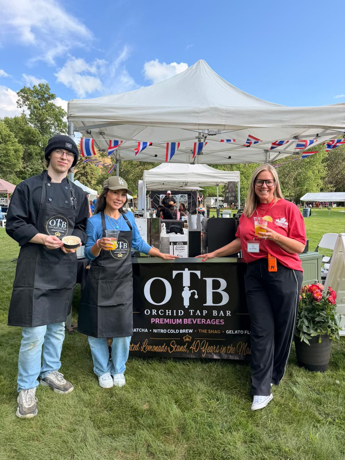 Three members of the OTB team in aprons holding drinks in front of the booth under a blue sky